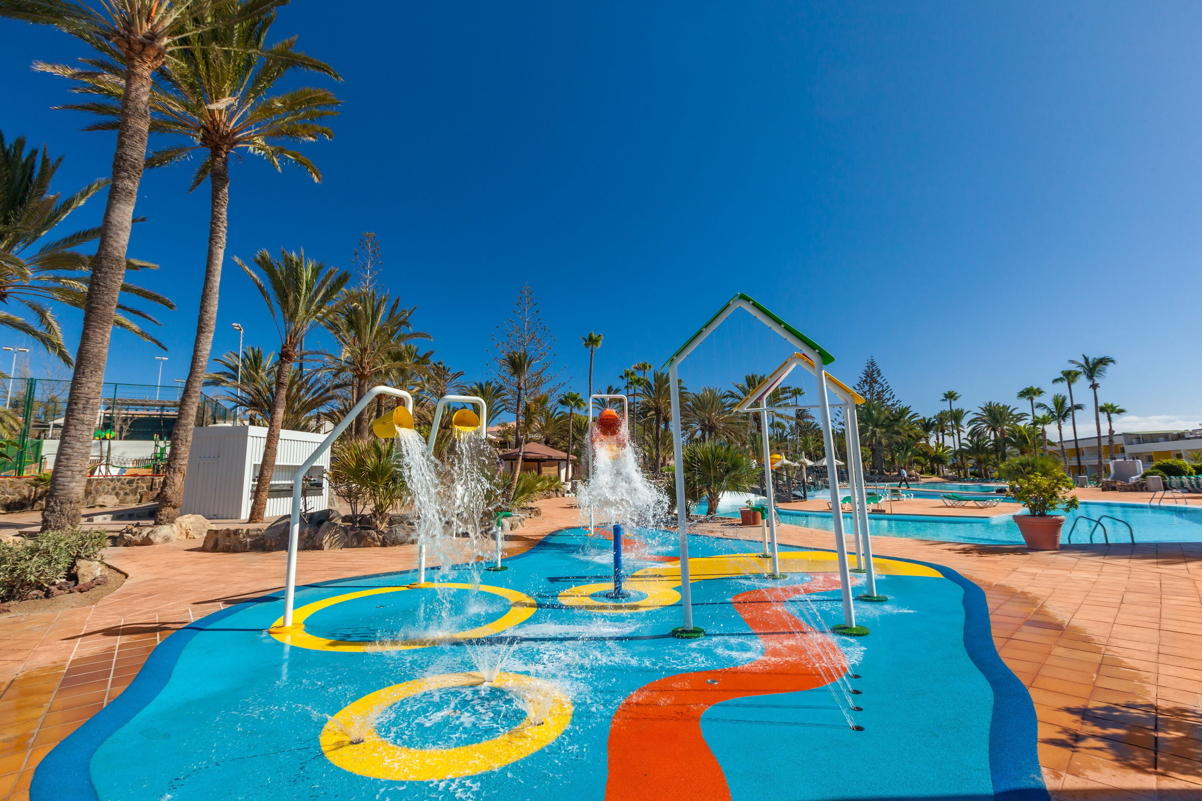 A water play area with multiple water jets and colorful ground markings, surrounded by palm trees and a clear blue sky.