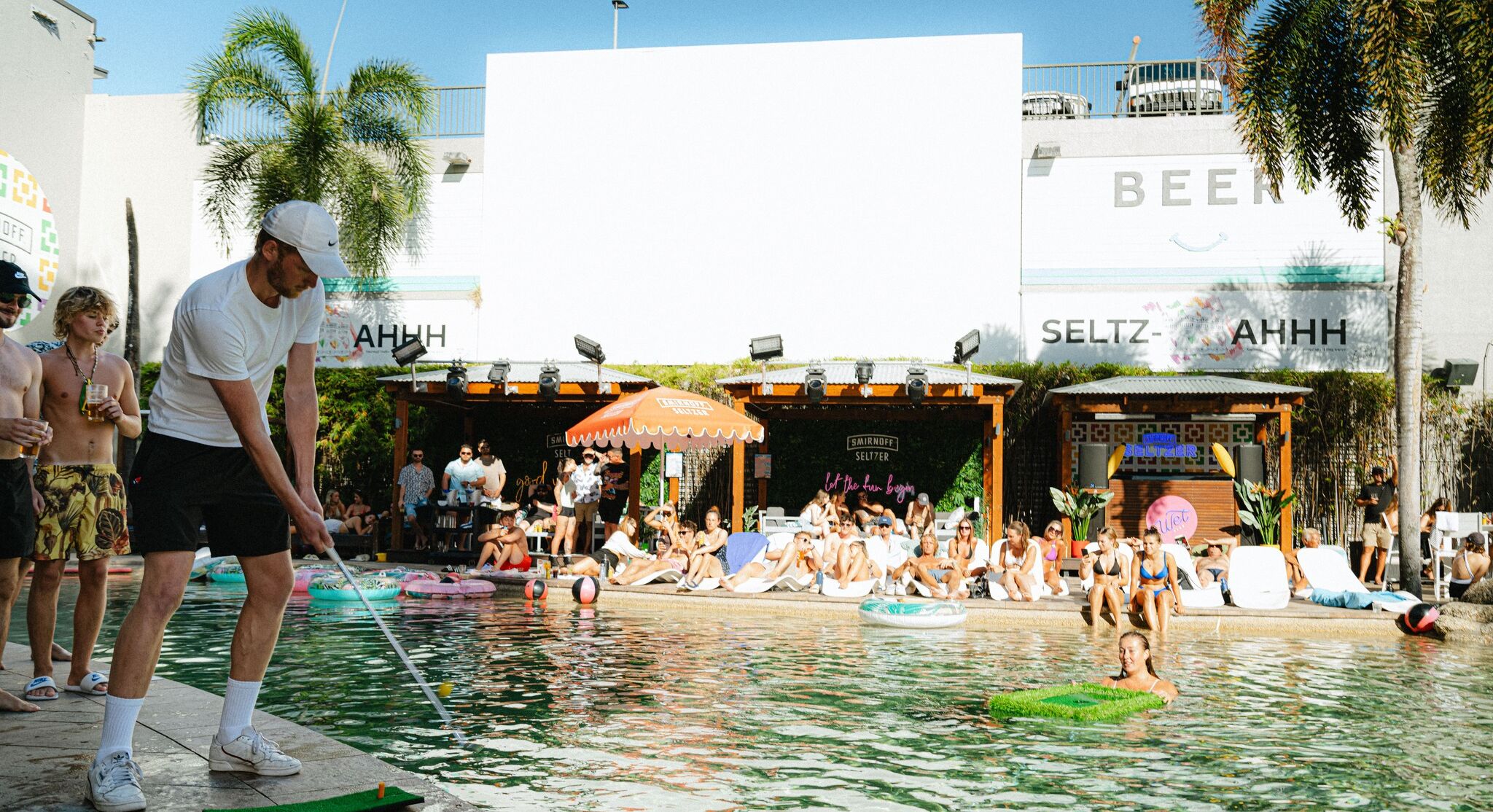 A pool area with lounge chairs and umbrellas, surrounded by palm trees and buildings with signage.