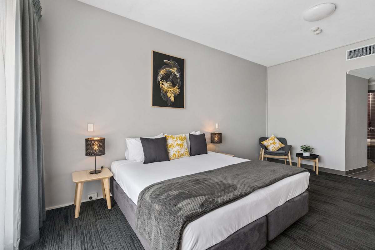 A modern hotel room with a large bed, two bedside tables with lamps, a small chair, and a decorative wall piece.