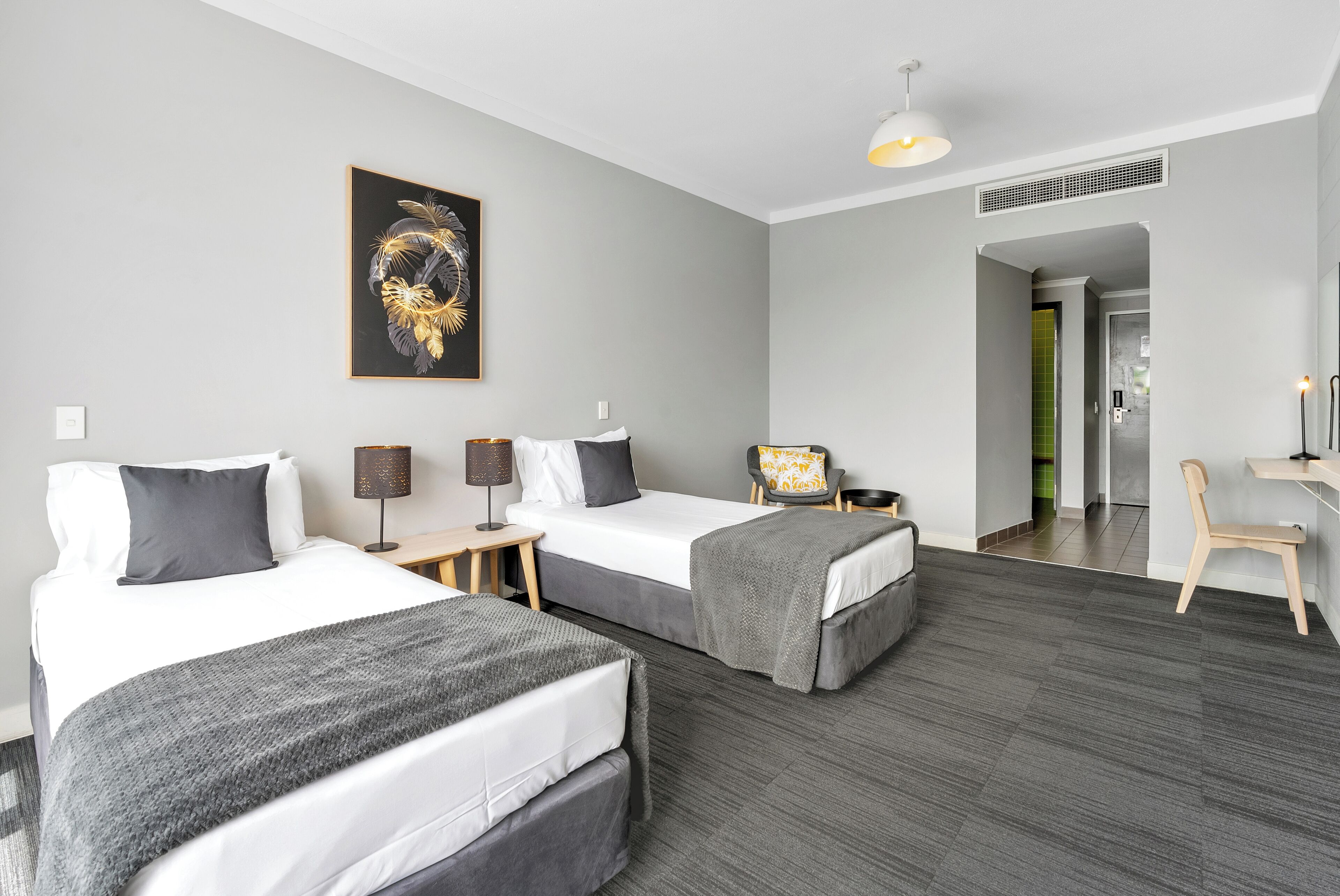 A hotel room with two beds, a desk, a chair, a lamp, and a framed artwork on the wall.