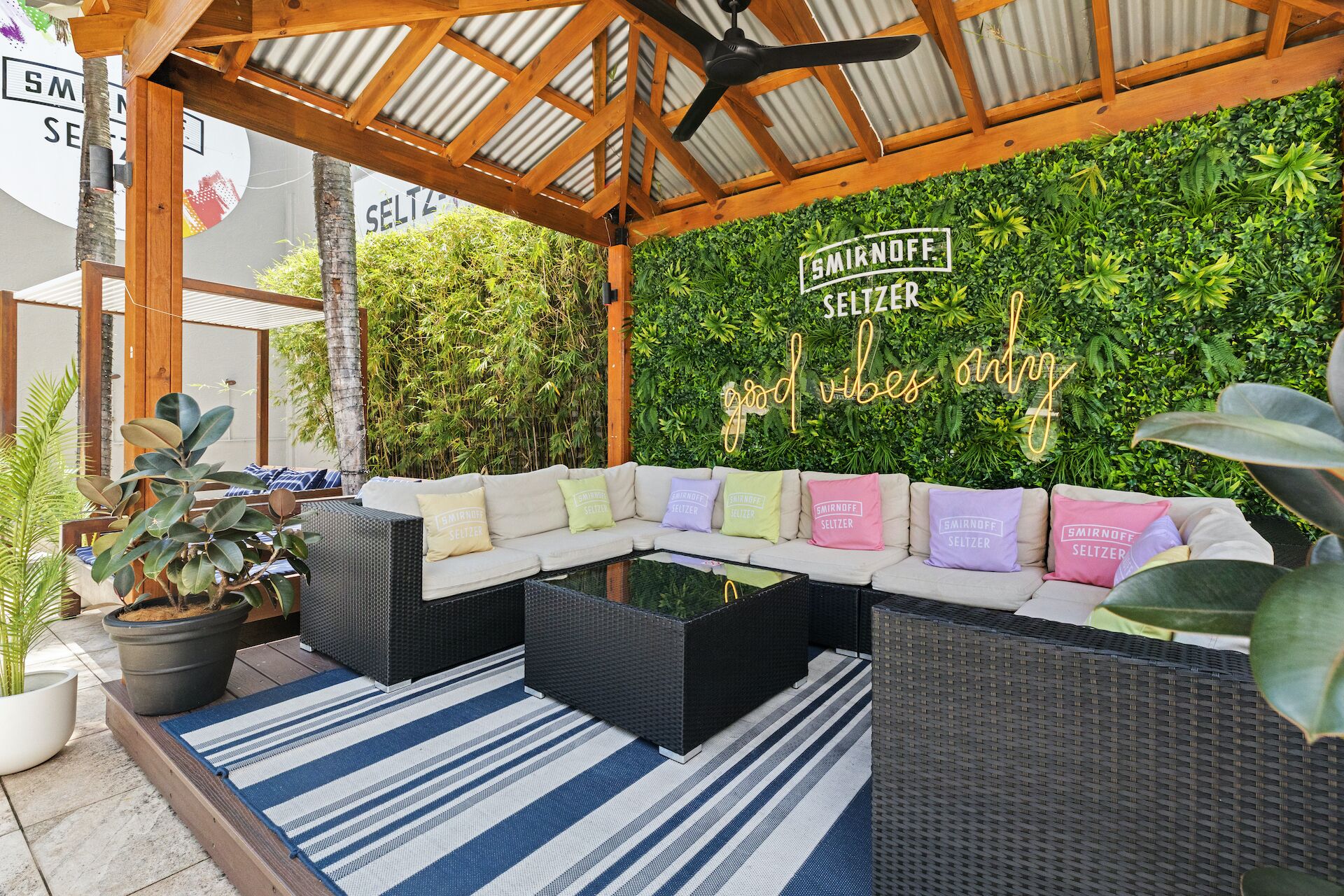 An outdoor seating area with a black wicker sofa, a white sofa with colorful cushions, and a green wall with a sign reading "SMIRNOFF SELTZER good vibes only".
