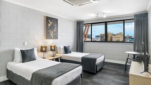 A hotel room with two beds, a desk, a chair, a lamp, and a large window with a city view.