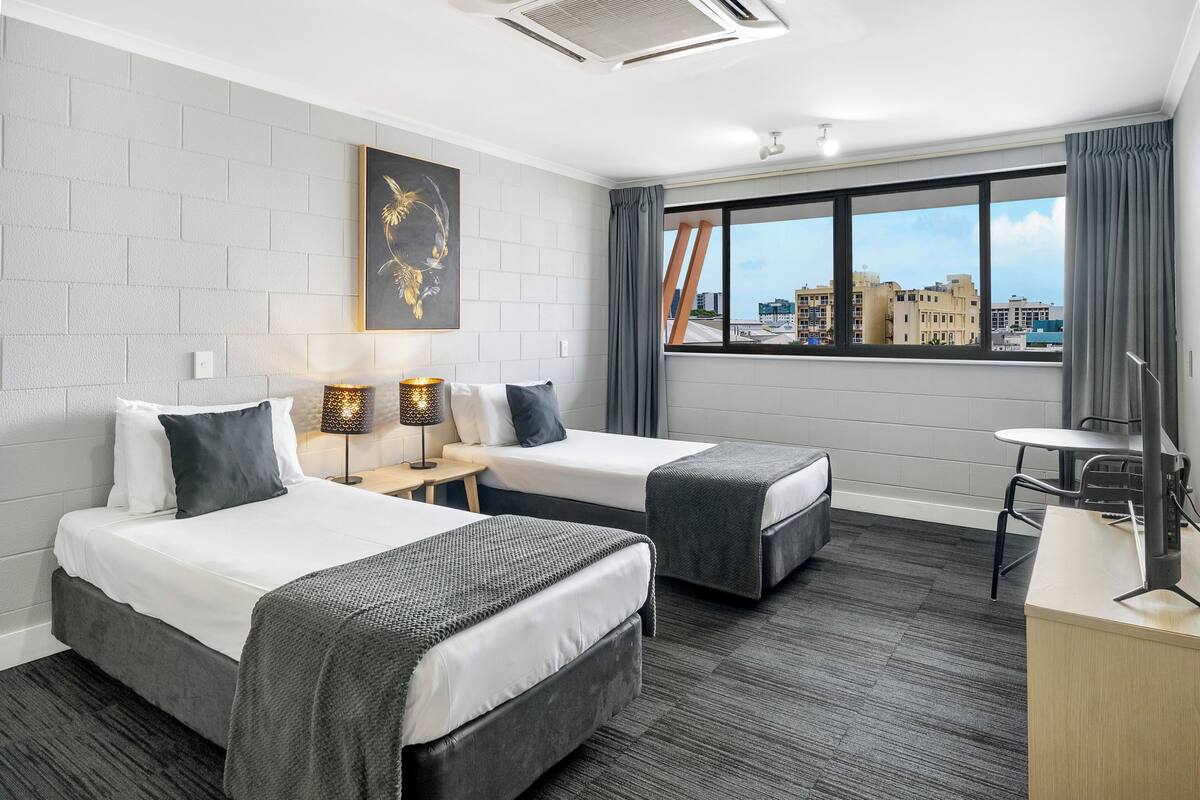 A hotel room with two beds, a desk, a chair, a lamp, and a large window with a city view.