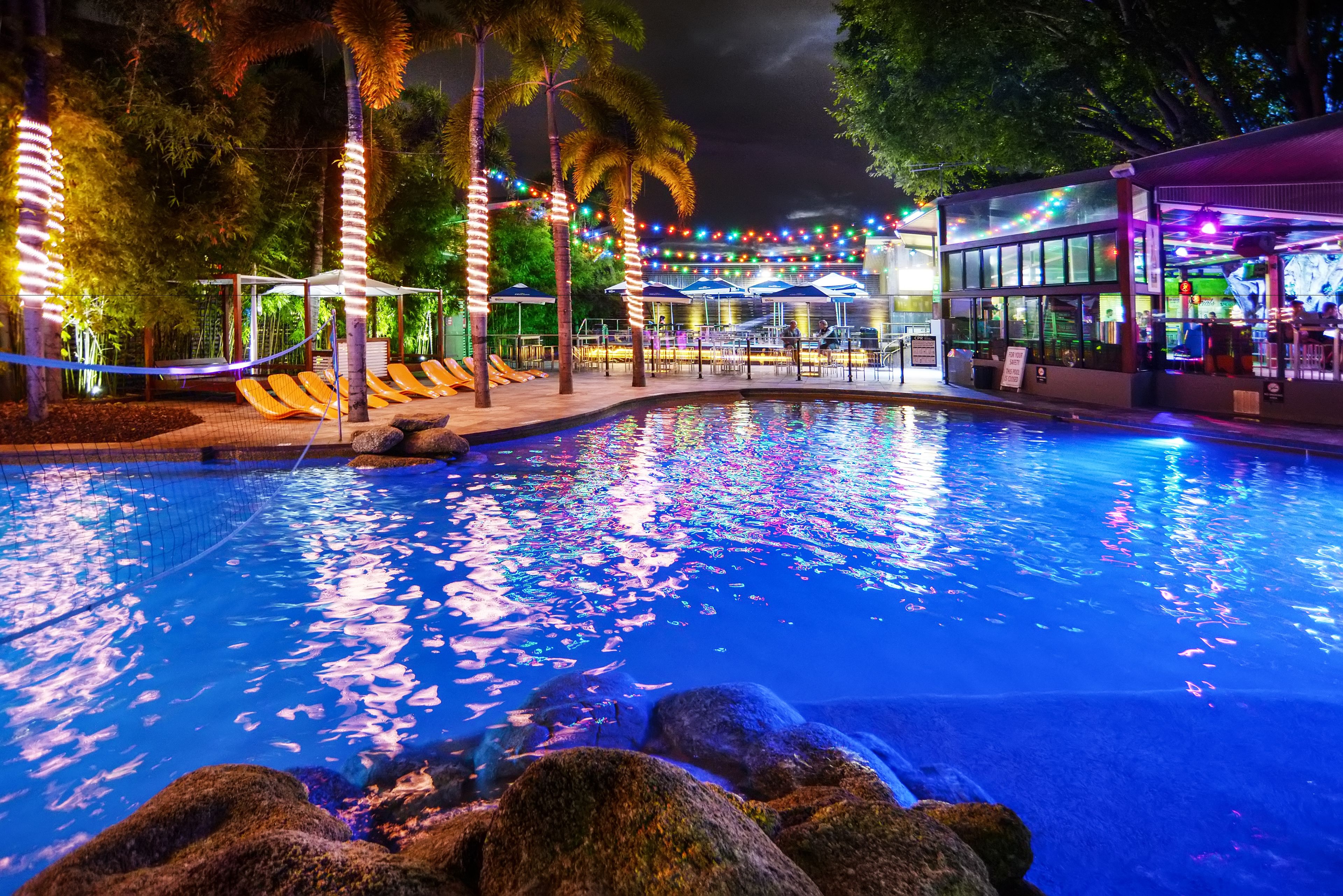 Un área de piscina nocturna con fuente de agua, sillas de descanso y una zona de estar cubierta.