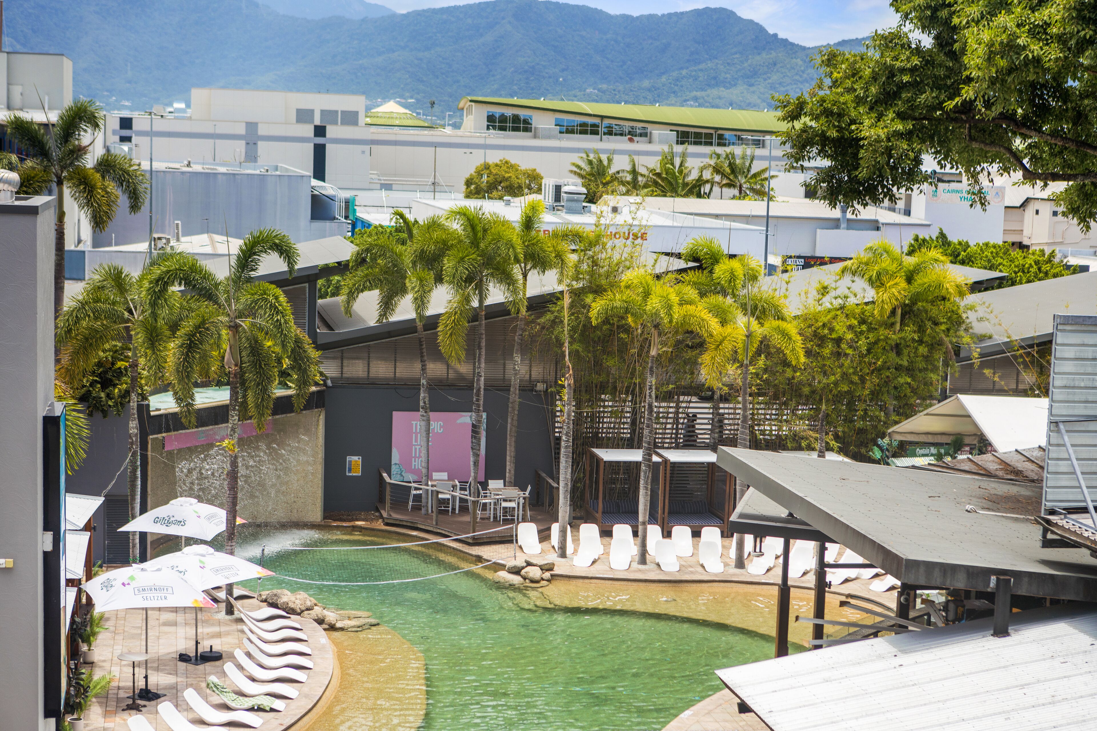 A pool area with lounge chairs and umbrellas, surrounded by palm trees and modern buildings.
