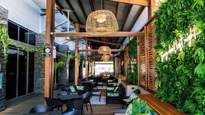 An indoor patio area with wooden tables and chairs, a hanging wicker light fixture, and a wall covered in green foliage.