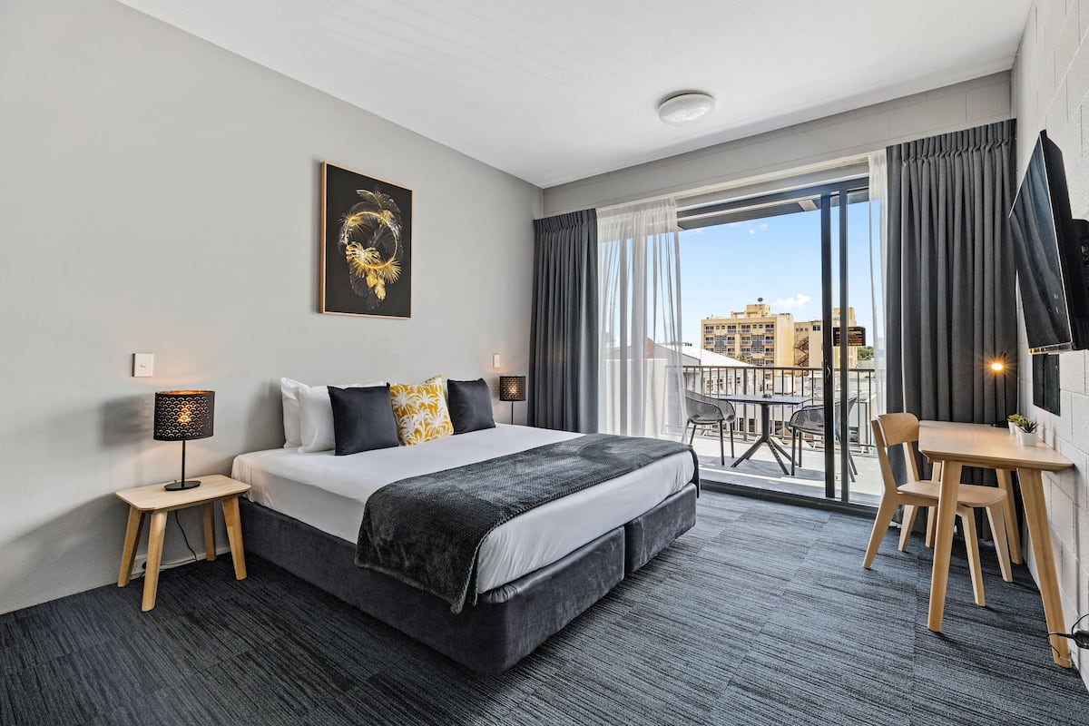 A modern hotel room with a large bed, a small table, a chair, and a balcony with a view of the city.