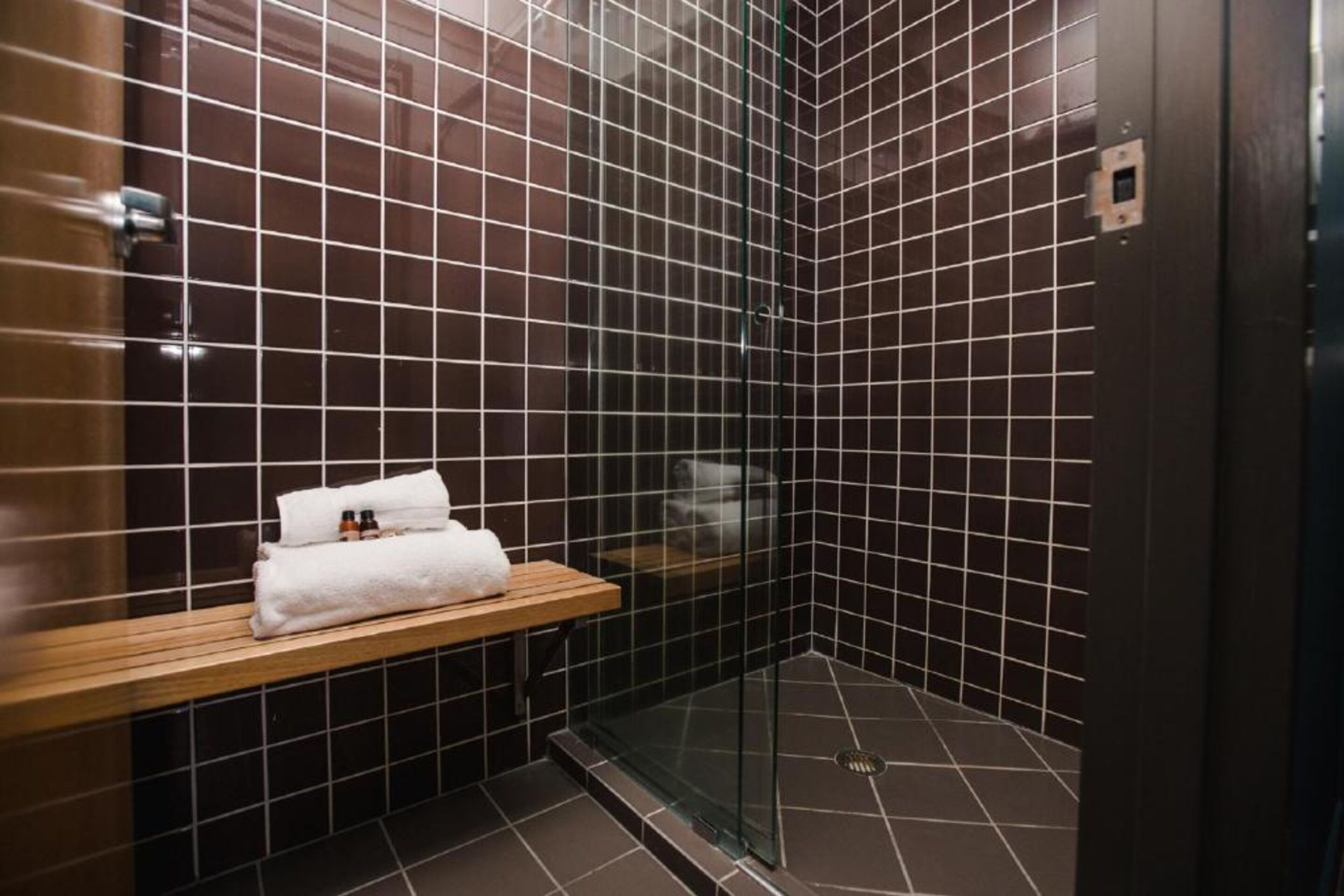 Une salle de bain moderne avec des murs recouverts de carreaux foncés, une douche en verre et une étagère en bois sur laquelle sont posés une serviette et des articles de toilette.