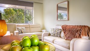 A modern living room with a sofa, a coffee table with green apples, and a large window with a view of trees. - Birches Serviced Apartments (East Melbourne)
