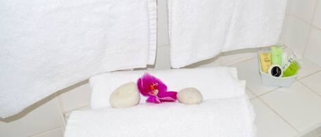 Un baño con dos toallas blancas colgadas en un perchero, un tapete de baño blanco, una orquídea rosada y un pequeño recipiente blanco con artículos de aseo.