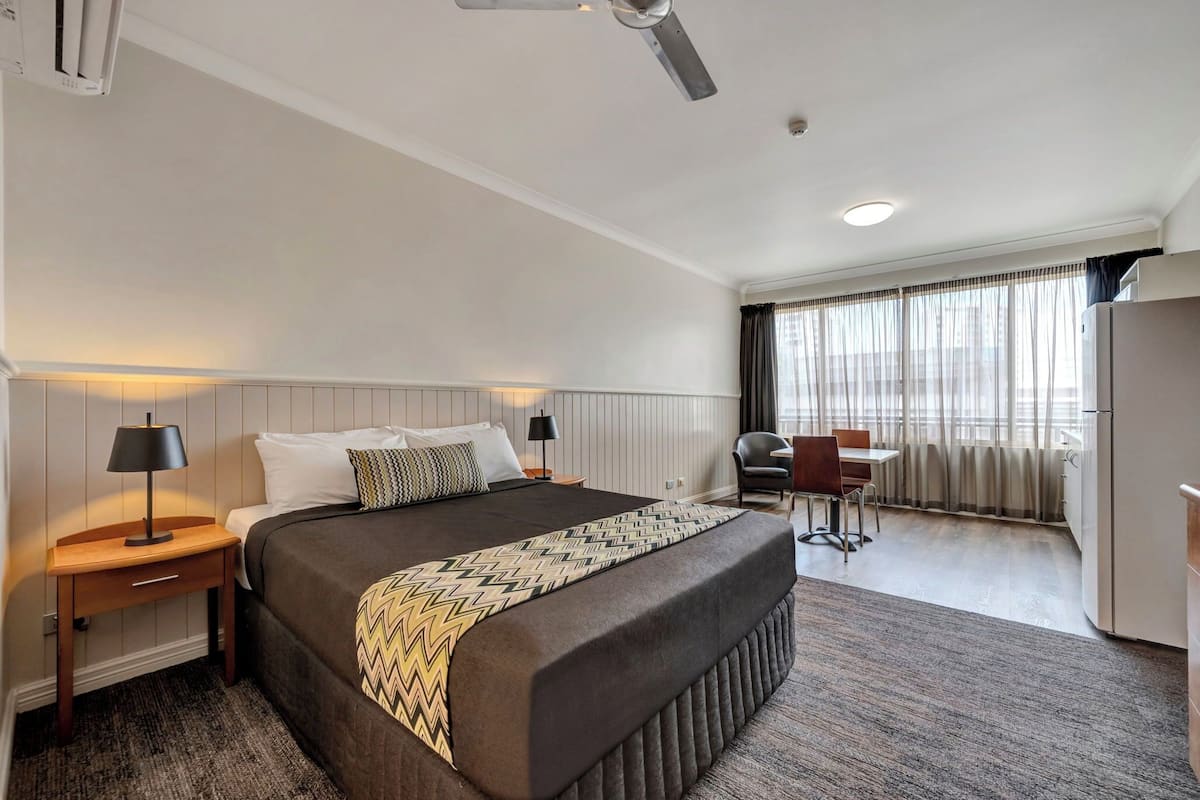 Une chambre d’hôtel avec un grand lit, des tables de chevet avec des lampes, un bureau avec une chaise, un réfrigérateur et une fenêtre avec des rideaux.