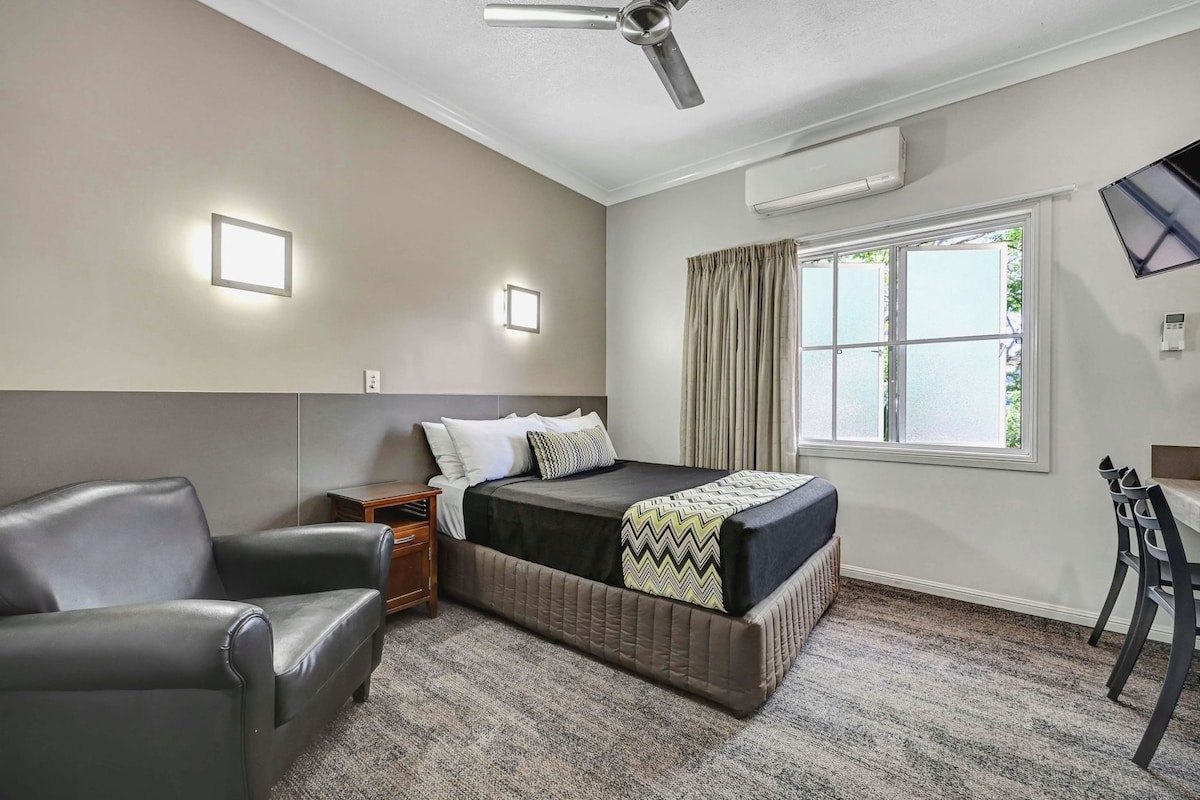 A hotel room with a bed, a chair, a bedside table, a window with curtains, an air conditioning unit, and a ceiling fan.