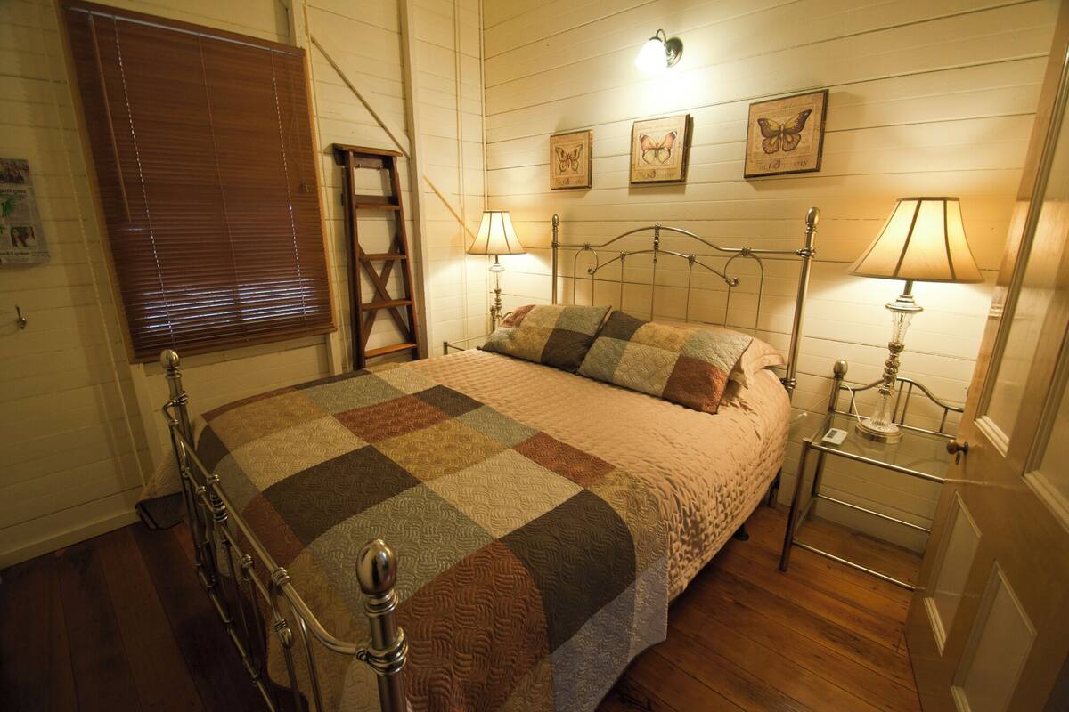 Une chambre avec une couvre-lit matelassé, deux lampes de chevet, un cadre de lit en métal, une échelle en bois et des œuvres d’art représentant des papillons accrochées au mur.