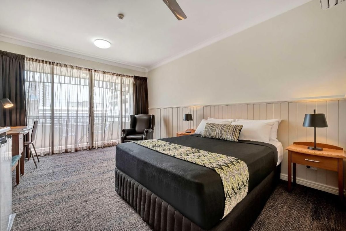 A hotel room with a large bed, a desk, a chair, and a window with curtains.