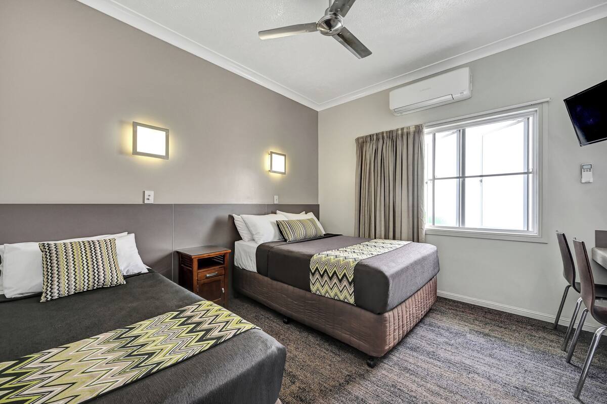 Une chambre d’hôtel avec deux lits, un ventilateur de plafond, un appareil de climatisation, une télévision et une fenêtre avec des rideaux.