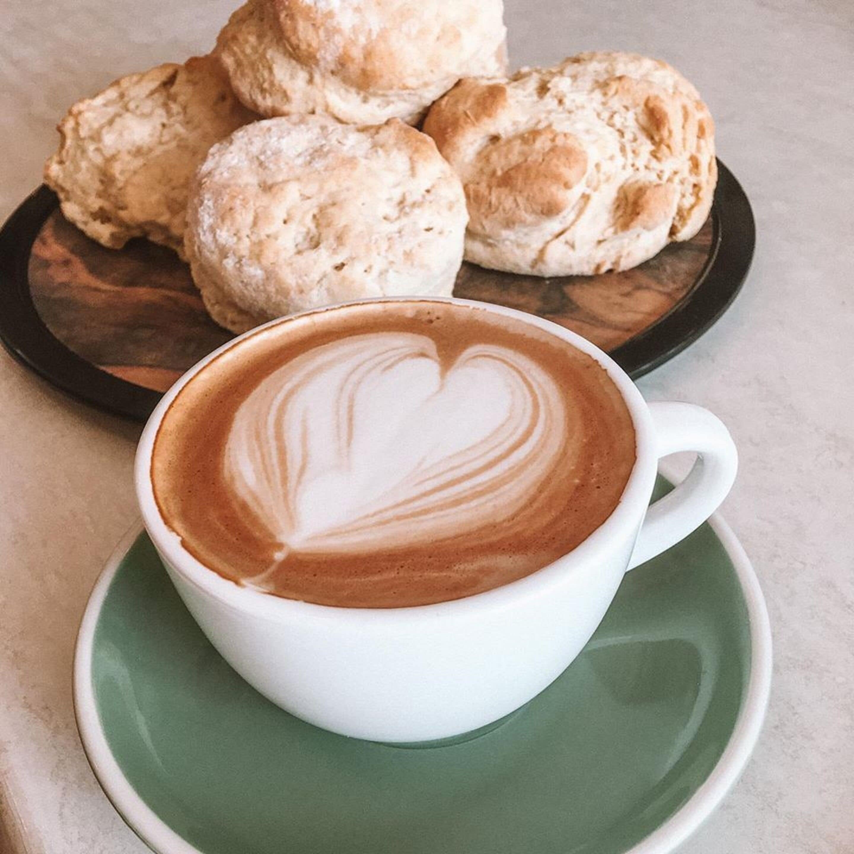 A cup of coffee with a heart-shaped latte art on a white saucer, accompanied by a plate of biscuits.