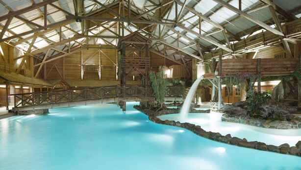 An indoor swimming pool with a wooden roof structure, a small waterfall feature, and a stone-lined edge. - Disney Davy Crockett Ranch (Bailly-Romainvilliers)