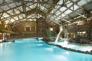 An indoor swimming pool with a wooden roof structure, a small waterfall feature, and a stone-lined edge.