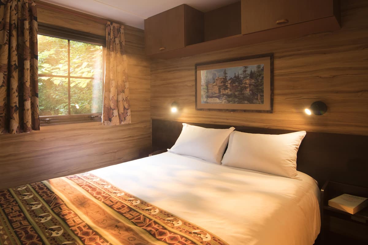 A neatly made bed with white linens, a patterned rug, a painting on the wall, and a window with curtains.