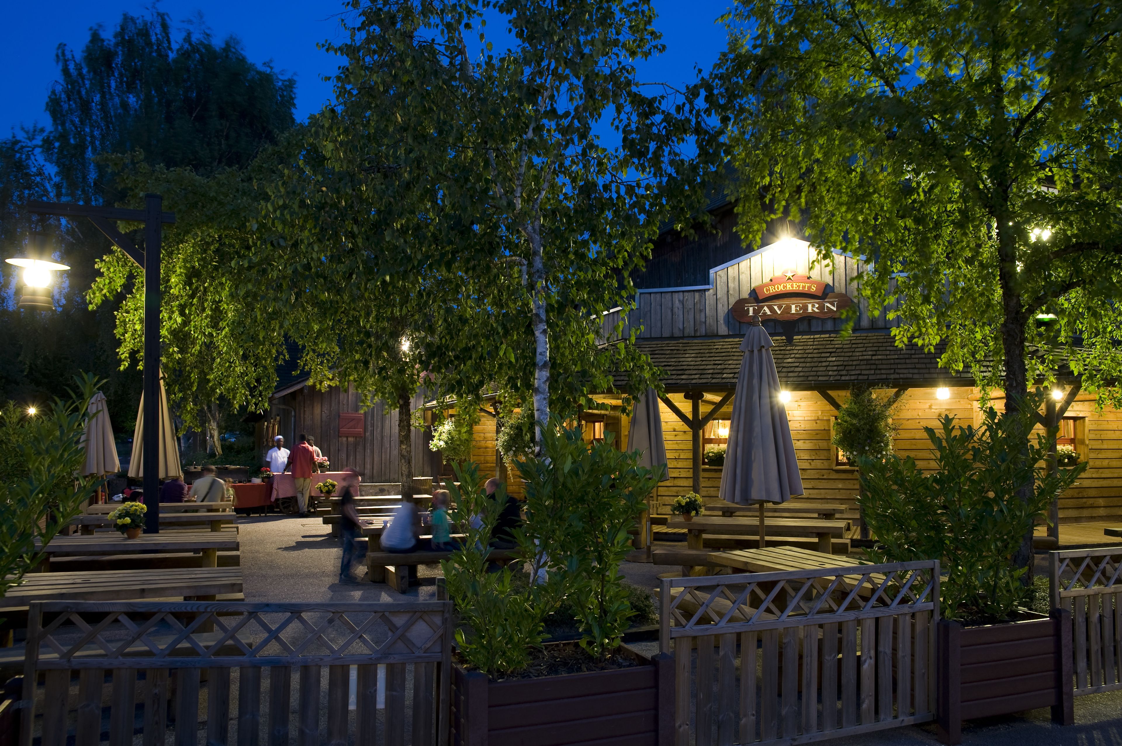 An outdoor seating area with wooden benches and tables, surrounded by trees and a wooden fence.
