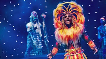 A performer in a colorful costume with a large headdress, standing on a stage with a dark background and spotlights.