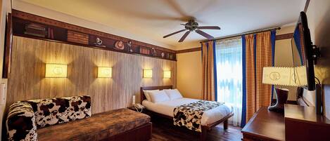 A hotel room with a wooden bench, a bed with a cowhide blanket, a ceiling fan, and a nightstand with a lamp.