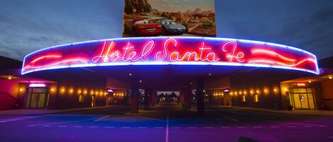 A hotel entrance with a neon sign reading "Hotel Santa Fe" and a large poster of animated cars above it.