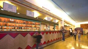 A reception area with a counter displaying various items, signs indicating arrival and departure, and a Disney Village sign in the background. - Disney Hotel Santa Fe (Coupvray)