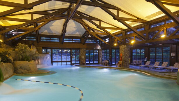 An indoor swimming pool with a waterfall feature, wooden beams, and lounge chairs. - Disney Sequoia Lodge (Coupvray)