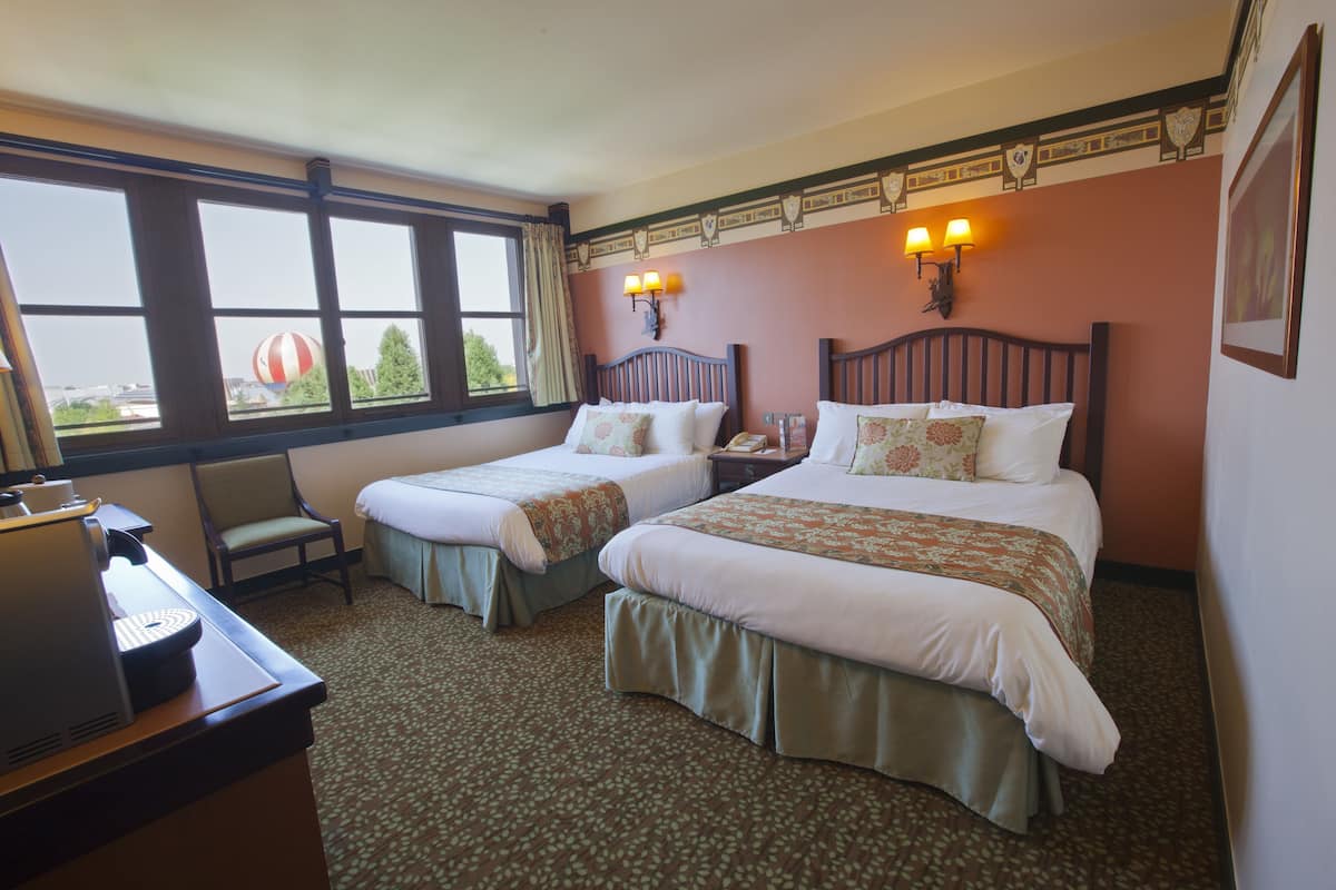 Habitación de hotel con dos camas, un escritorio con cafetera, una silla y ventana con vista a un globo aerostático.
