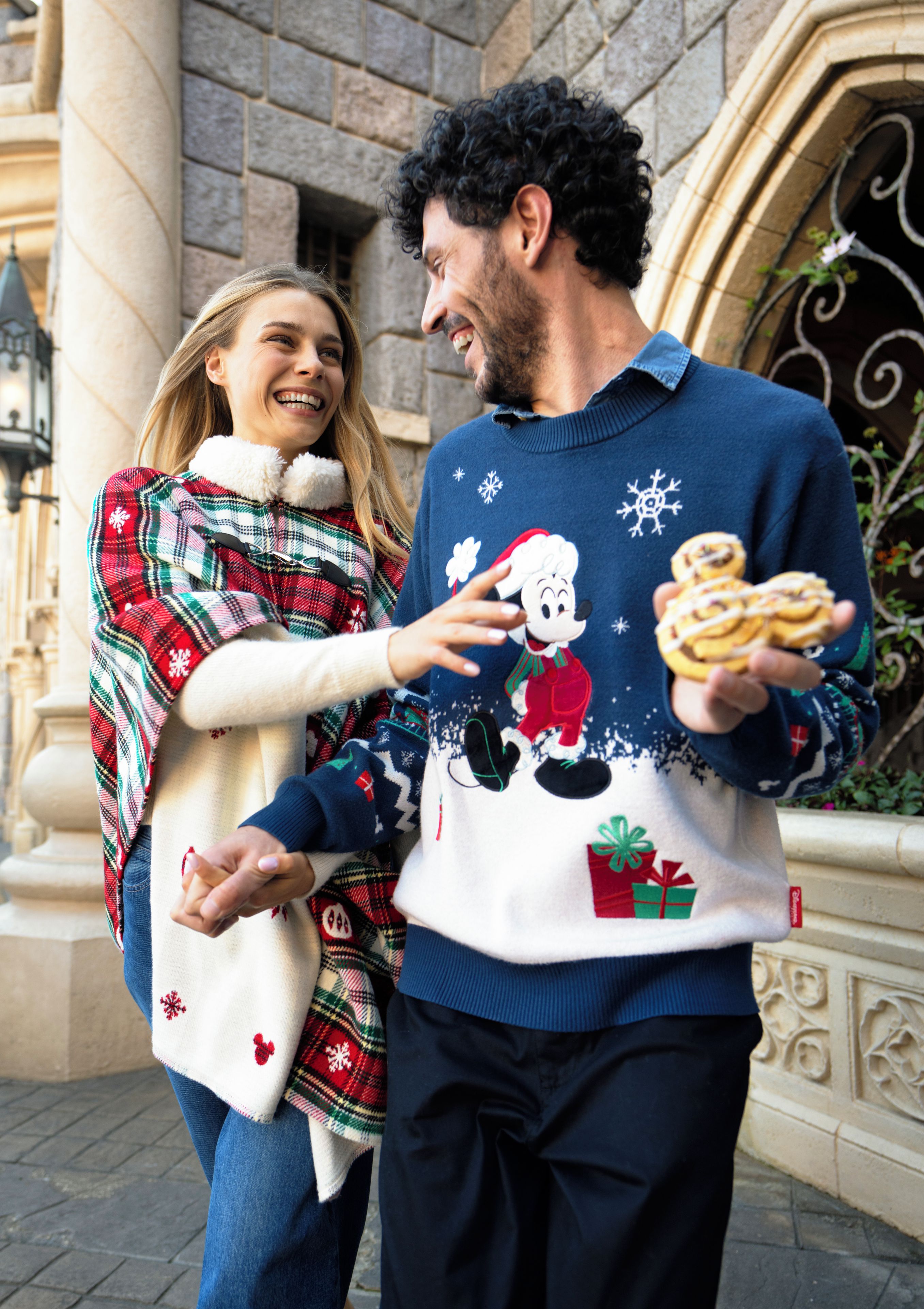 Une personne tenant une assiette de nourriture, portant un pull festif orné d’un motif de bonhomme de neige, et se tenant devant un bâtiment en pierre avec une porte en arc de cercle.