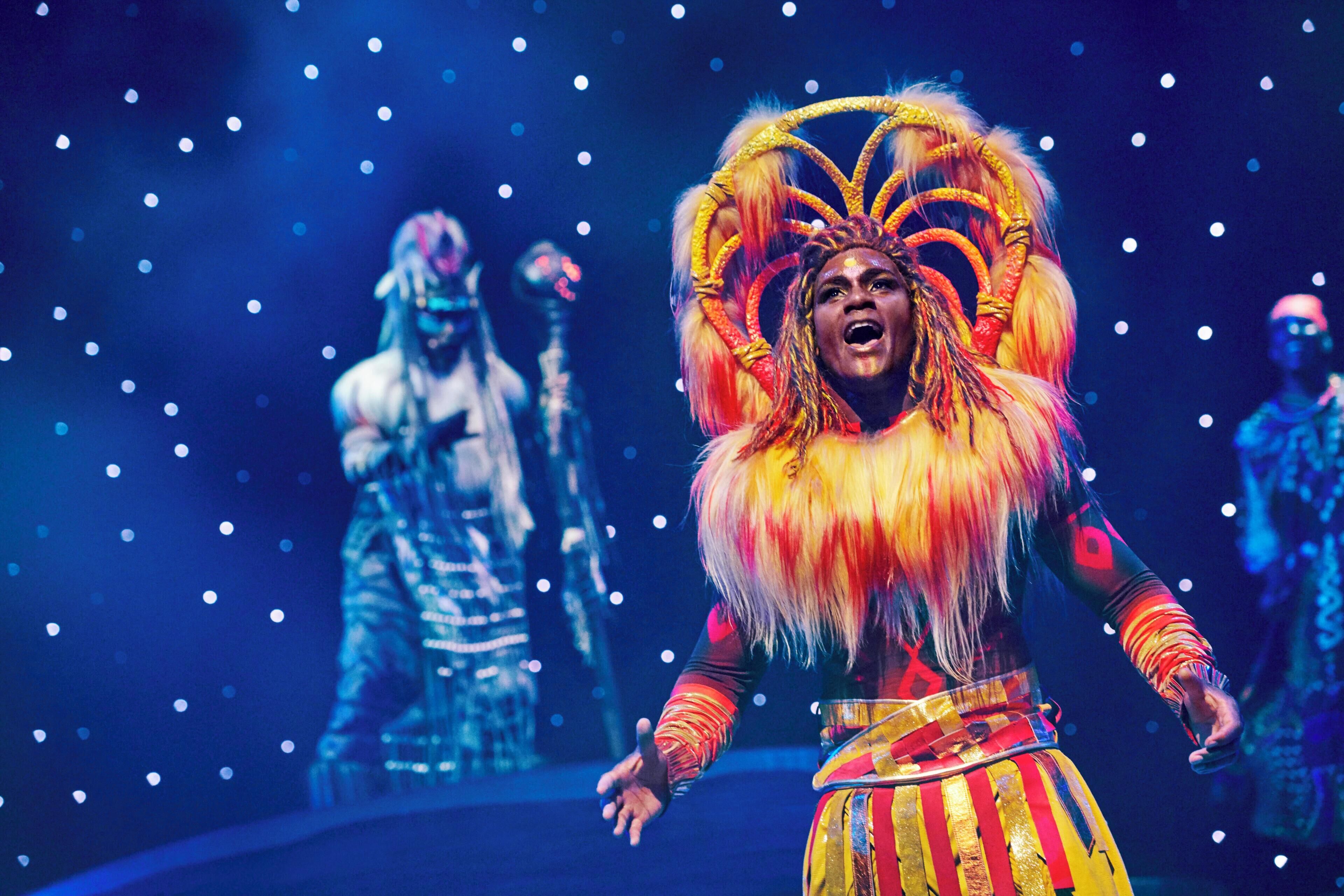 A performer in a colorful costume with a large headdress, standing on a stage with a dark background and spotlights.