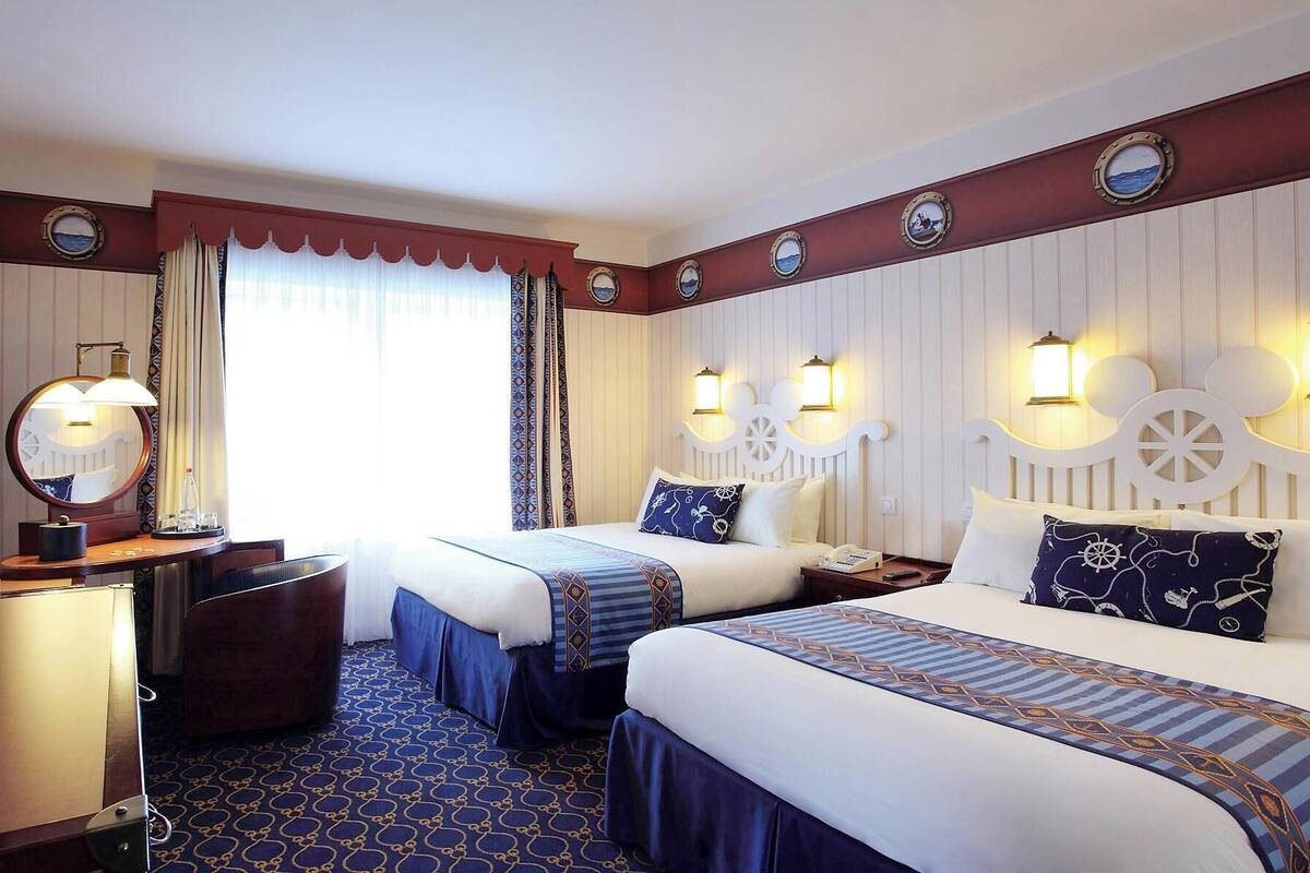 A hotel room with two beds, a desk with a mirror, a chair, and a large window with curtains.