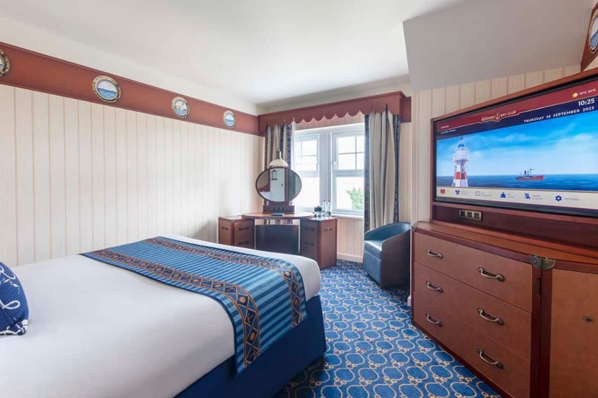 A hotel room with a bed, a desk, a dresser, and a television displaying a lighthouse scene.