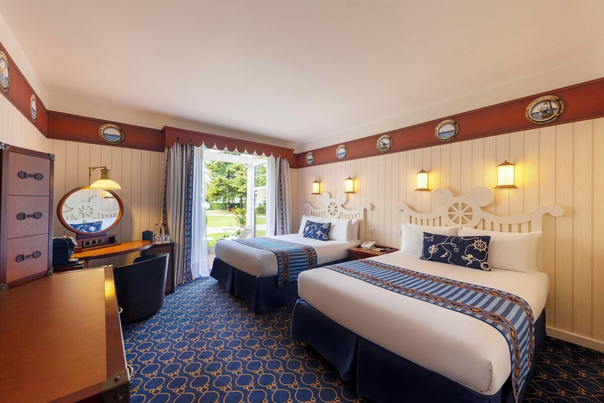 A hotel room with two beds, a desk, a chair, and a large window with curtains.