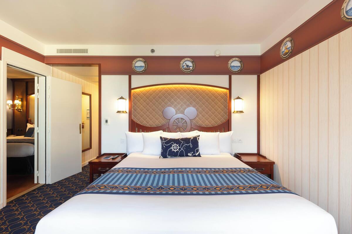 A hotel room with a large bed, a Mickey Mouse headboard, and a view of the hallway.