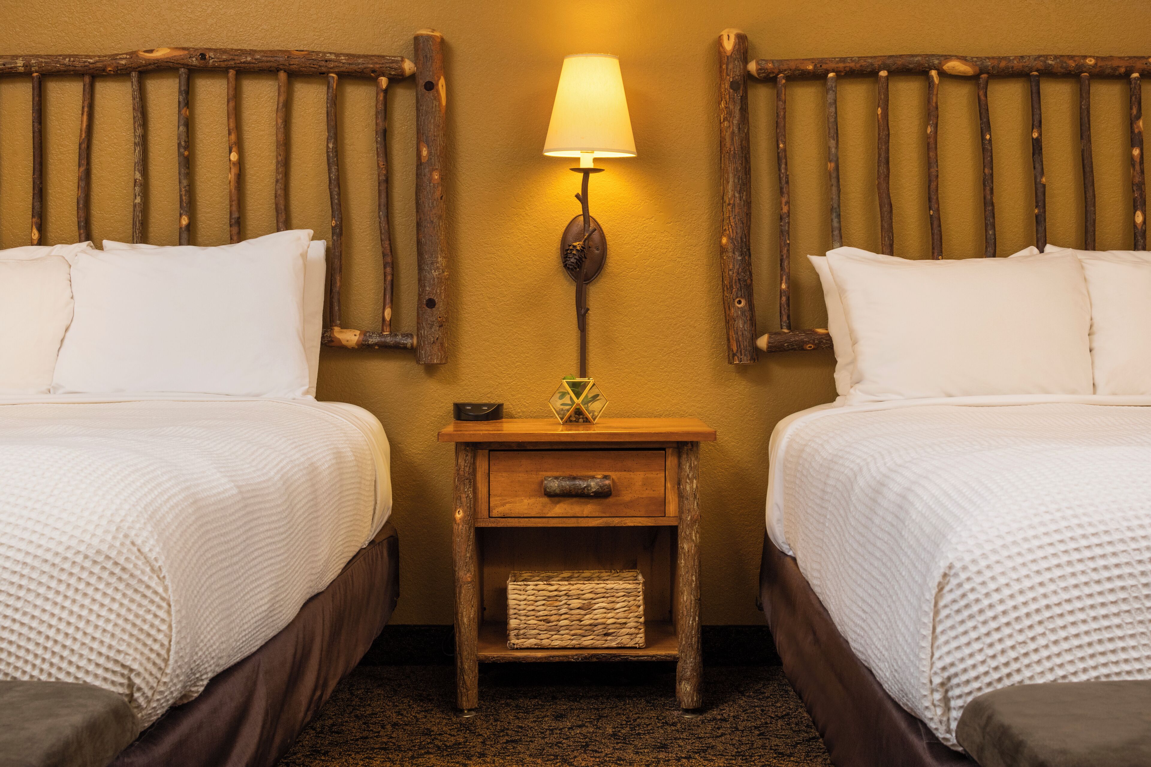 A hotel room with two beds, a nightstand with a lamp, and a woven basket on the nightstand.