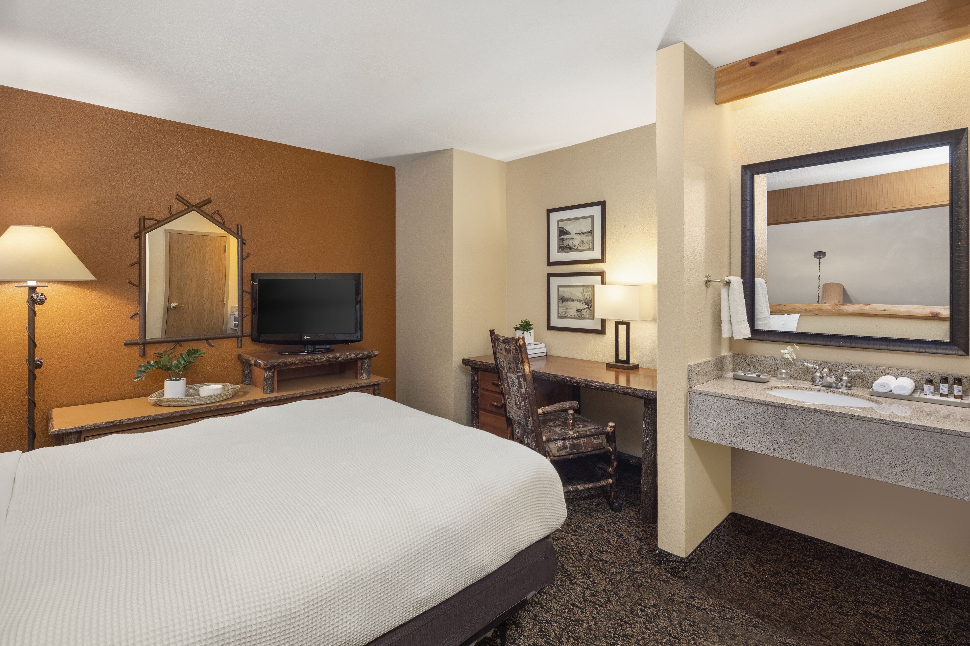 Een hotelkamer met een bed, bureau, stoel, televisie en een badkamer met een wastafel en spiegel.