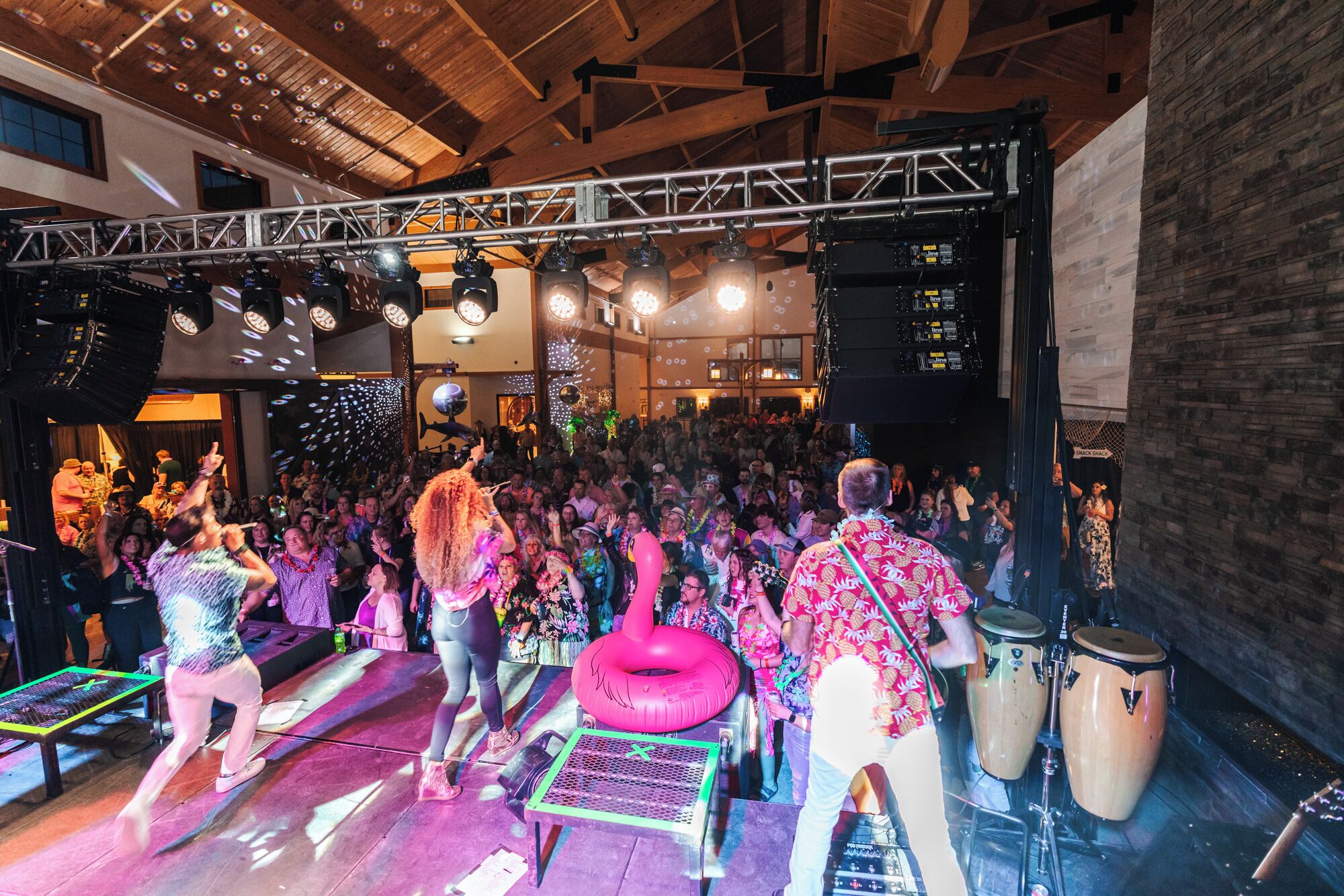 A crowded indoor event with a stage, DJ equipment, and a crowd of people.