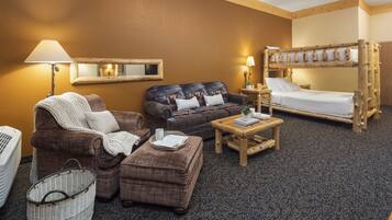 A cozy room with a sofa, armchair, ottoman, wooden bunk bed, and a small table with a lamp.