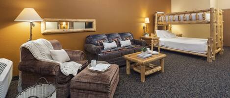 A cozy room with a sofa, armchair, ottoman, wooden bunk bed, and a small table with a lamp.