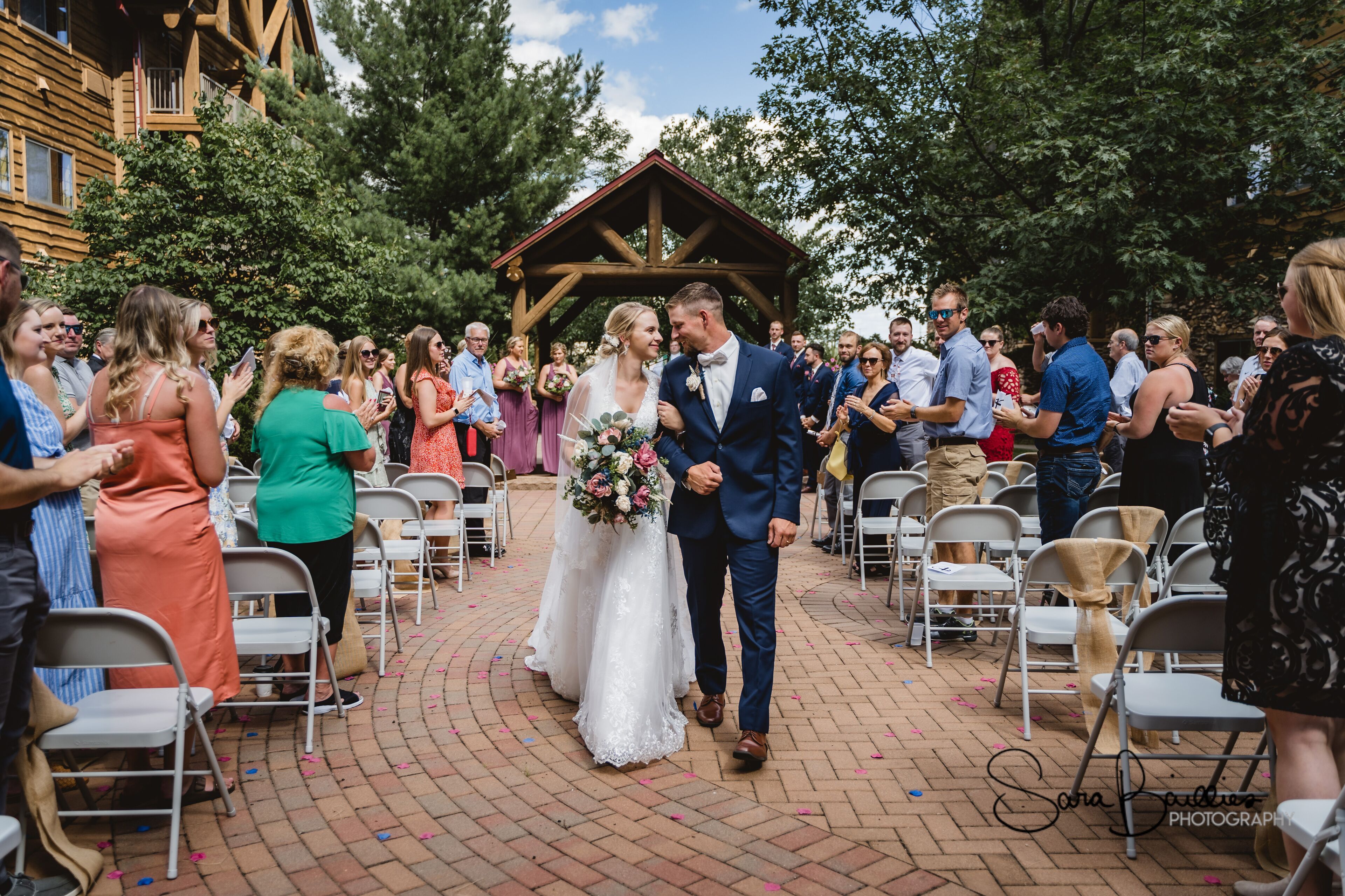 A wedding ceremony with guests seated on metal chairs arranged in rows, a wooden structure in the background, and a brick pathway.