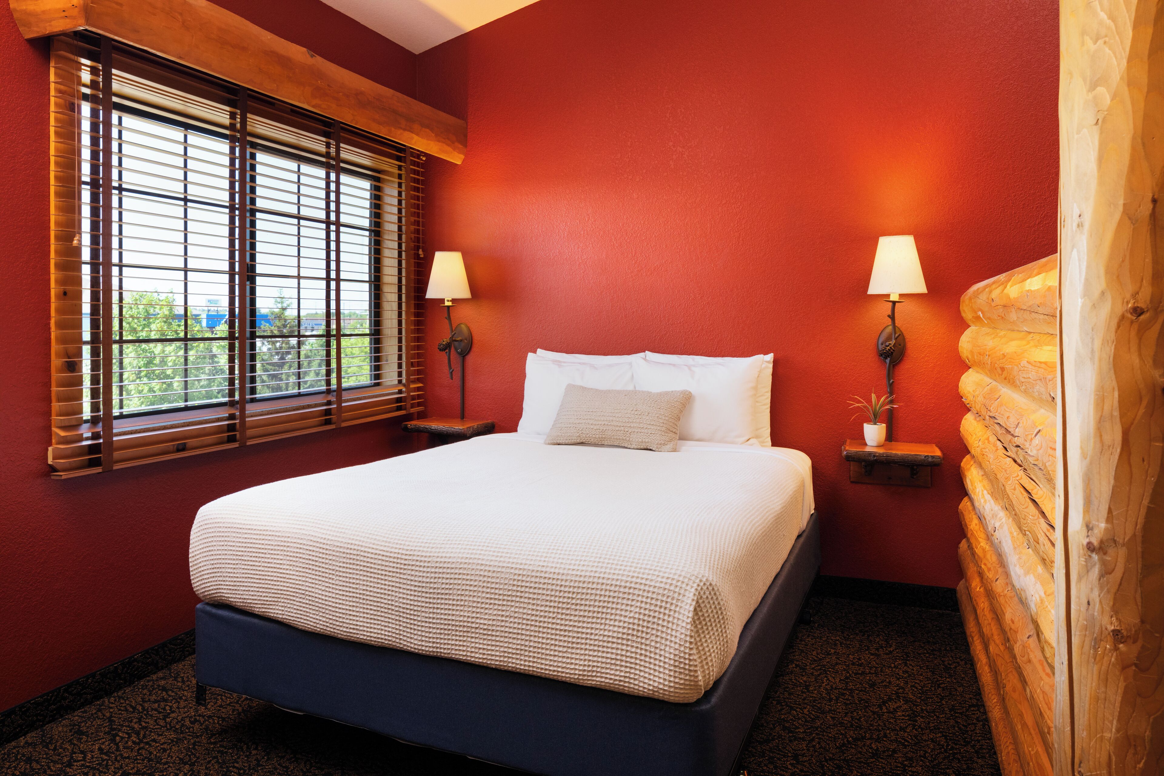 A hotel room with a large bed, two bedside lamps, a window with blinds, and a view of greenery outside.