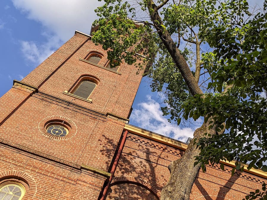 A brick tower with a clock and arched windows, surrounded by trees.
