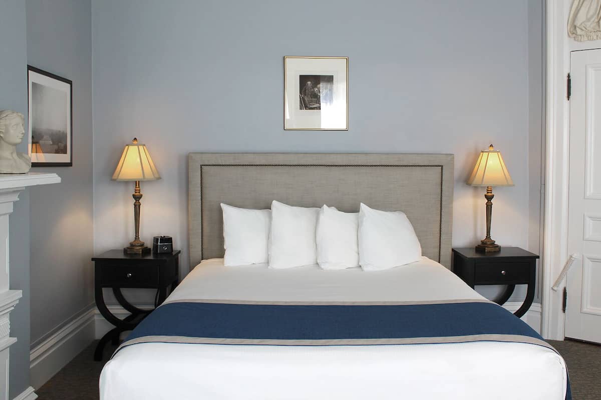 A neatly made bed with a grey headboard, white pillows, and a blue accent at the foot. There are two bedside tables with lamps, a framed picture above the bed, and a white door to the right.