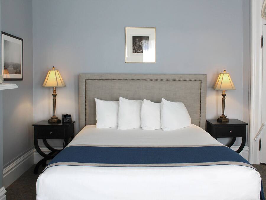 Una cama bien hecha con cabecera gris, almohadas blancas y un detalle azul al pie. Hay dos mesitas de noche con lámparas, un cuadro enmarcado sobre la cama y una puerta blanca a la derecha.