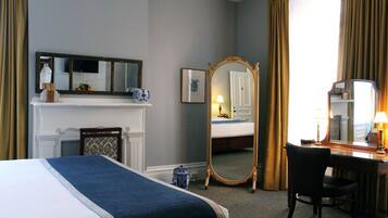 A hotel room with a bed, a desk, a chair, a dresser, a mirror, a fireplace, and a window with curtains.