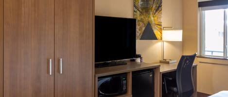A hotel room with a wooden wardrobe, a flat-screen TV, a microwave, a refrigerator, a desk with a lamp, and a chair.