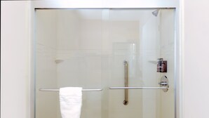A glass-enclosed shower with a white towel hanging on a metal bar, a wooden floor, and a small shelf with toiletries. - Boulder University Inn (Boulder)