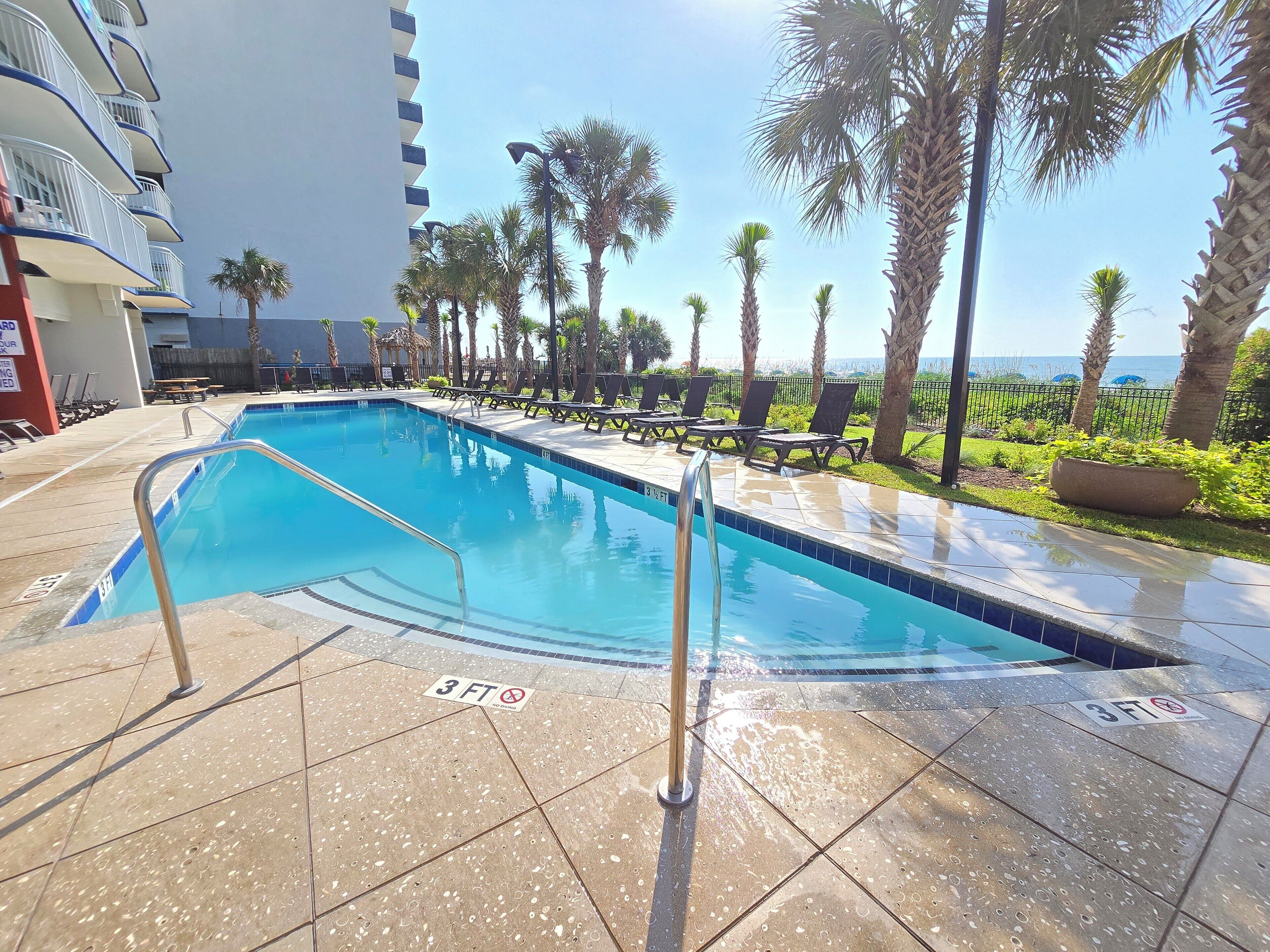 A swimming pool with a 3 ft depth marker, surrounded by lounge chairs and palm trees, with a building in the background.
